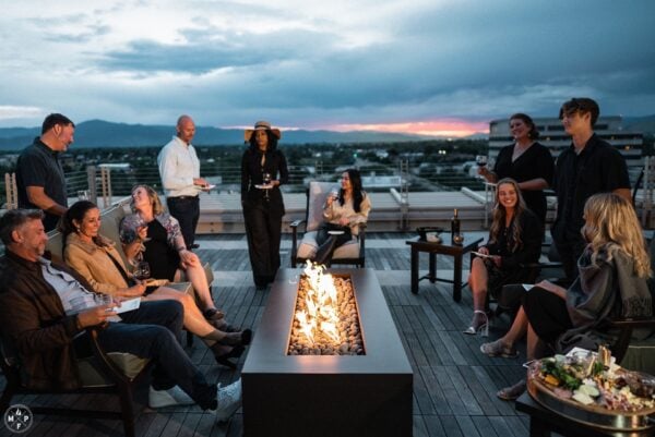group of people at a rooftop party around a venture series steel fire table called the olympic