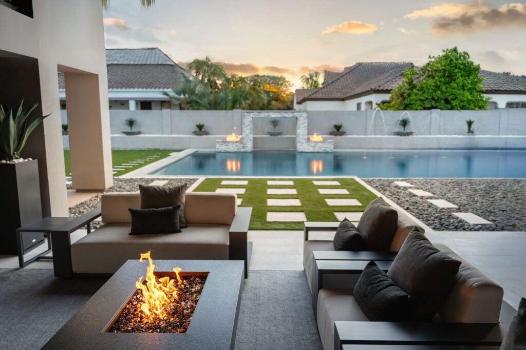 A picture of backyard design with a fire feature and some seating areas, a pool, an two built in fire features in the background.